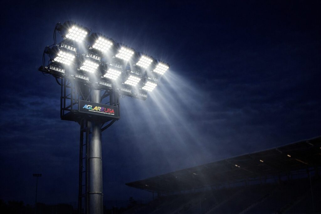 High-mast LED stadium flood lights casting bright white beams against a night sky at a sports venue